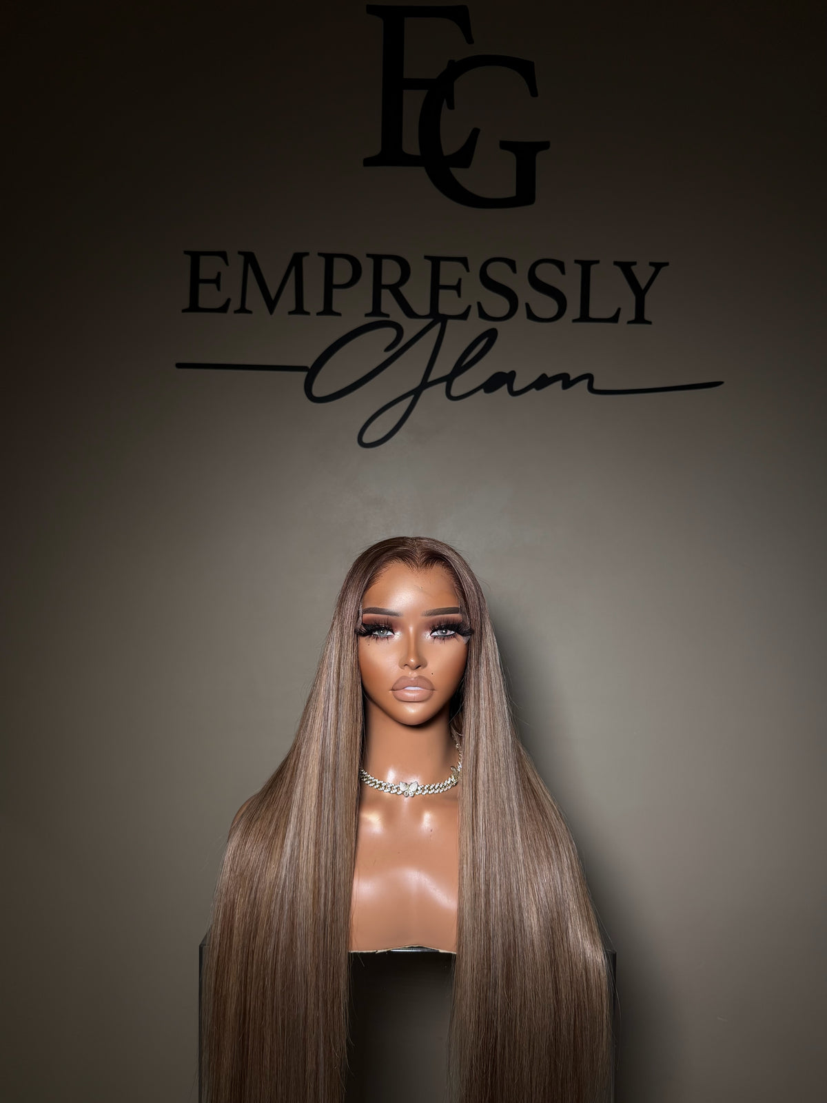 A long, straight wig displayed on a mannequin bust against a dark gray background, with the brand name 'Empressly Glam' and an 'EG' logo printed above. The wig has a warm brown root with smooth, highlighted ash-brown lengths.